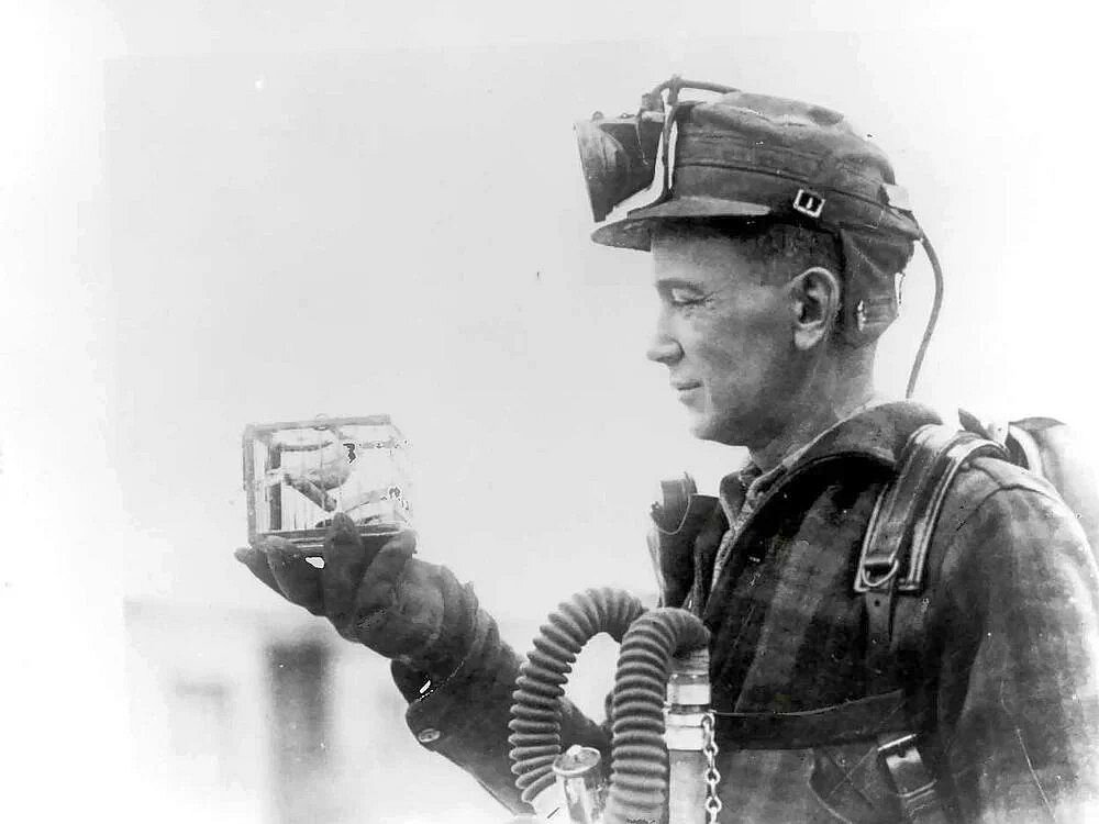 A coal miner in breathing apparatus cradles a small caged canary in his gloved hand, studying it